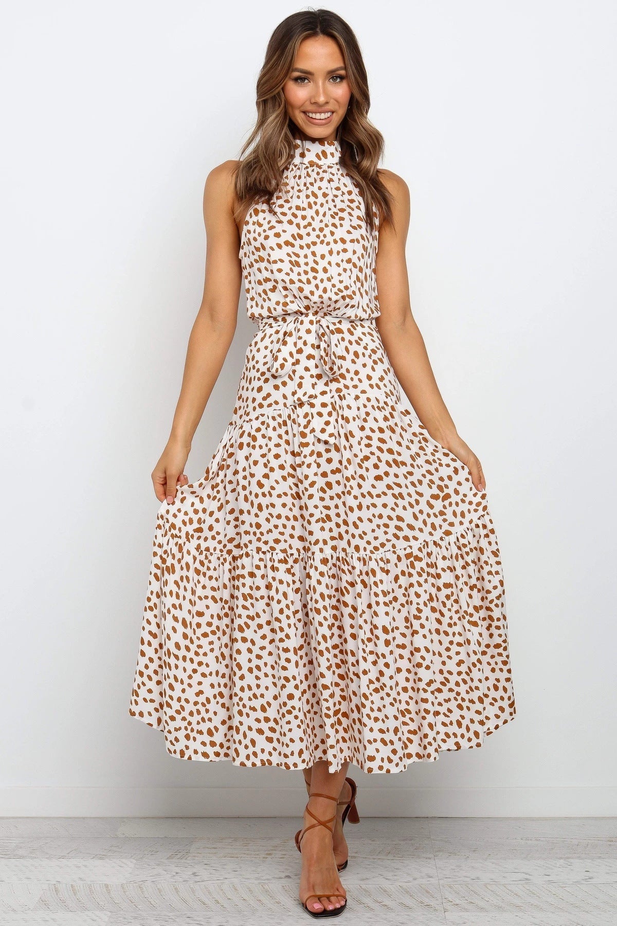 Frau in ärmellosem, geblümtem Maxikleid, beige mit braunen Punkten, posiert lächelnd vor weißem Hintergrund. Elegantes Sommeroutfit, Mode.