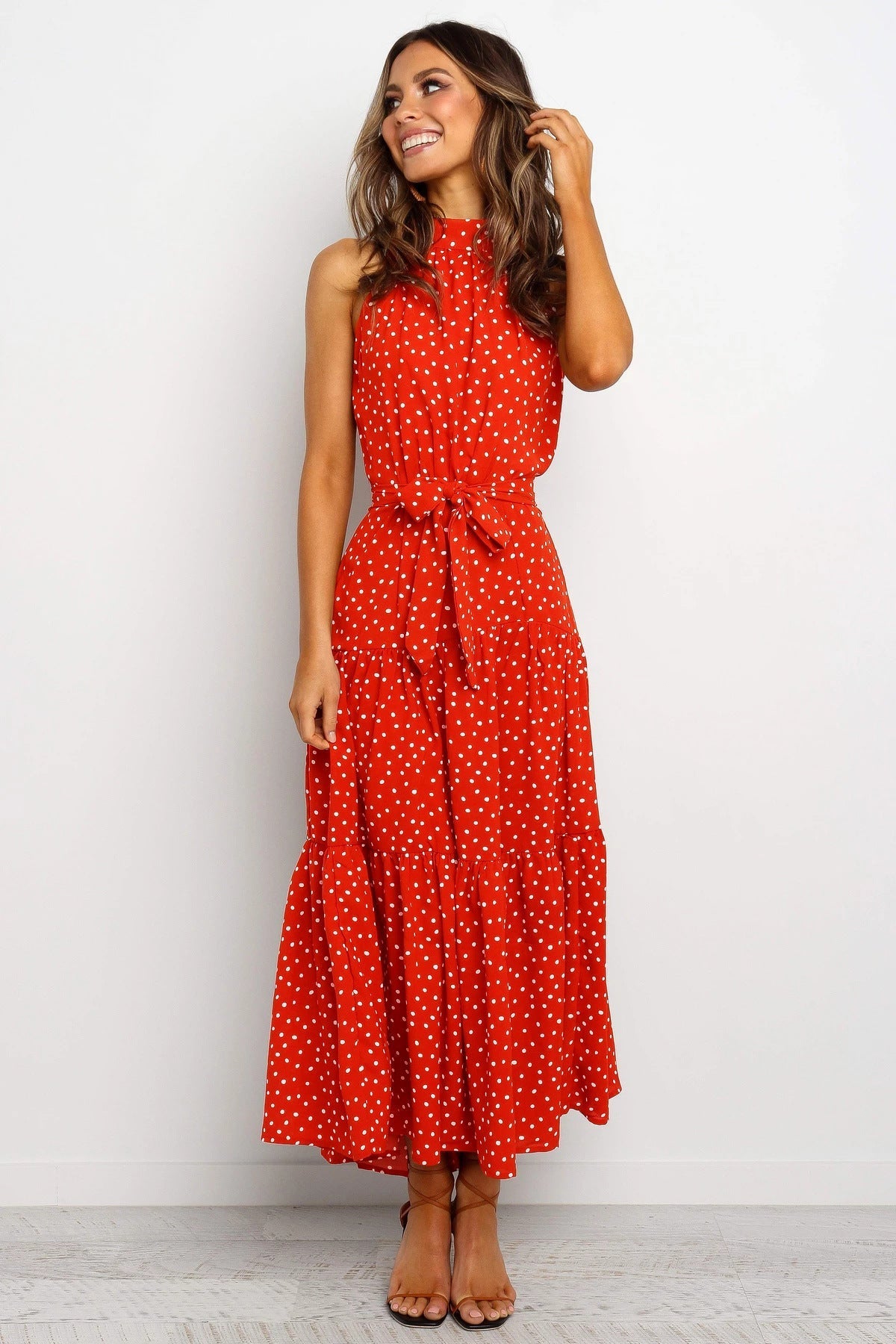 Frau in rotem, ärmellosem Maxikleid mit weißen Punkten, lächelt und steht vor weißem Hintergrund. Sommermode, elegantes Design, Freizeitkleidung.