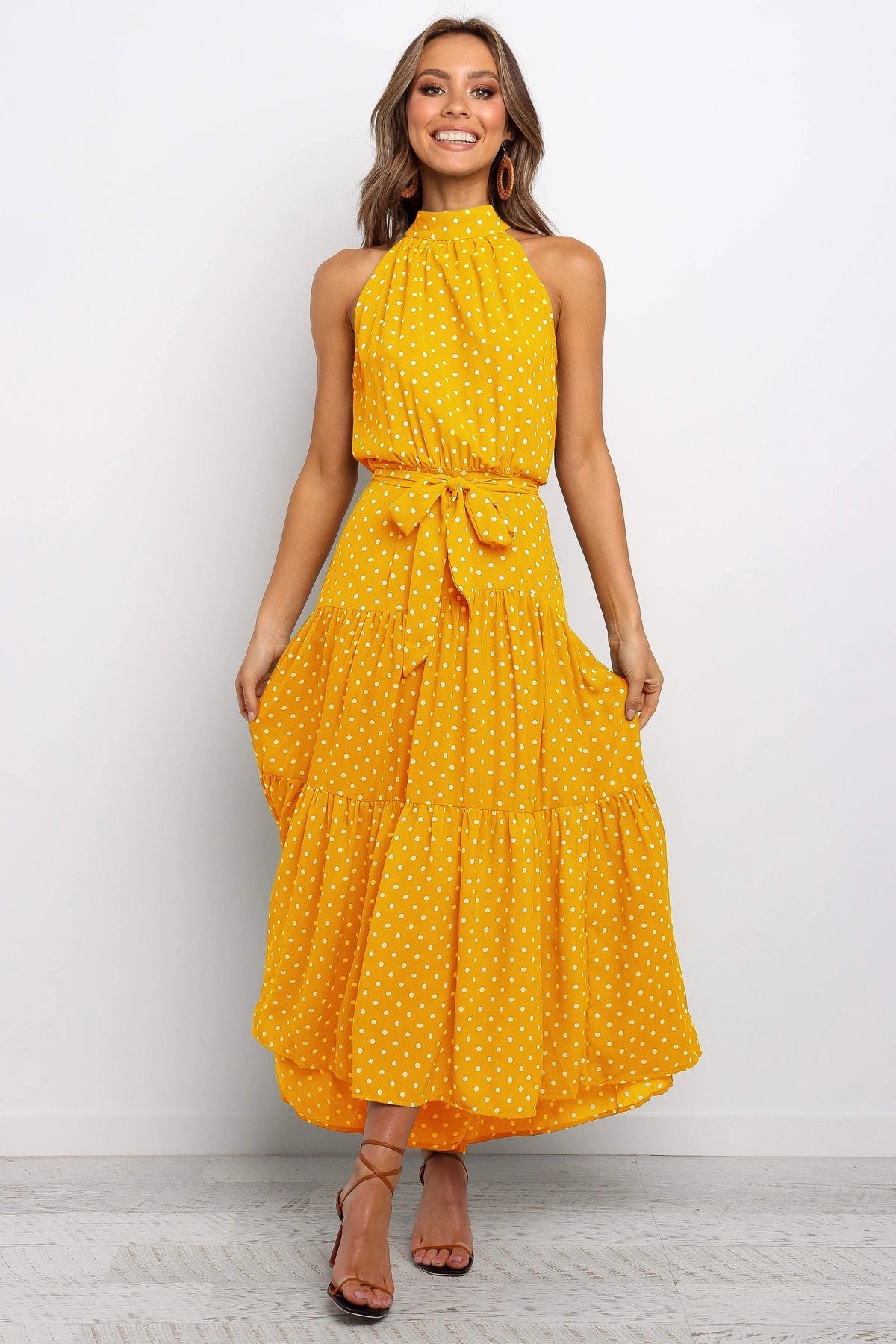 Frau in gelbem, gepunktetem Sommerkleid mit Halterneck und Taillengürtel, lächelnd, vor weißem Hintergrund. Elegantes, modisches Damenoutfit.