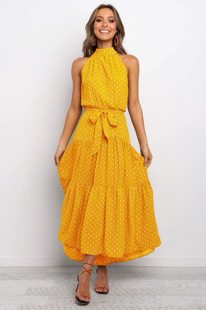 Frau in gelbem, gepunktetem Sommerkleid mit Halterneck und Taillengürtel, lächelnd, vor weißem Hintergrund. Elegantes, modisches Damenoutfit.