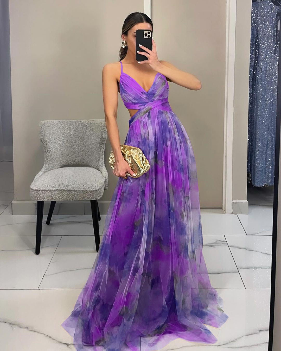 Frau in lila Abendkleid mit Blumenmuster, hält goldene Clutch, macht Selfie im Spiegel. Elegantes Outfit, Mode, Abendgarderobe.