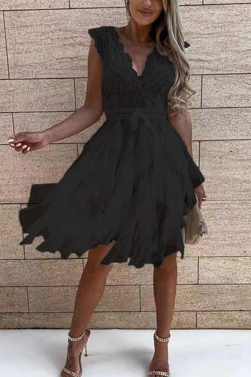 Frau in elegantem, schwarzem Spitzenkleid mit V-Ausschnitt und Rüschenärmeln, hält eine kleine Tasche, steht vor einer Ziegelwand.