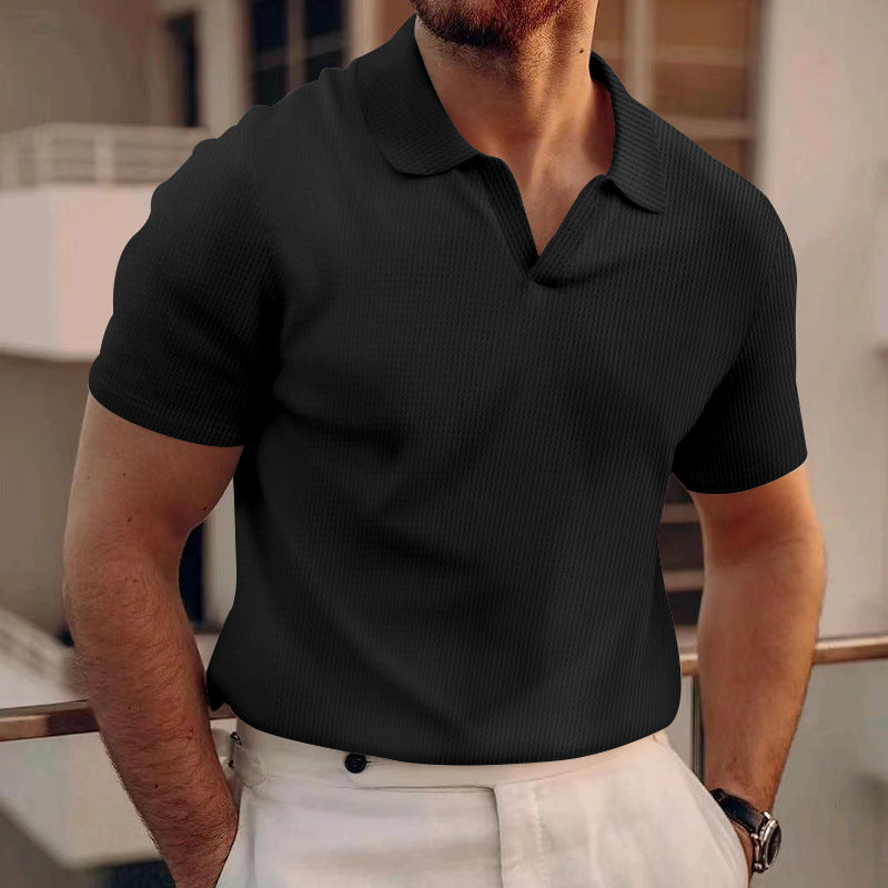 Mann in schwarzem Polohemd und weißer Hose auf Balkon, moderne Architektur im Hintergrund. Elegante Herrenmode, stilvolles Outfit, urbane Umgebung.