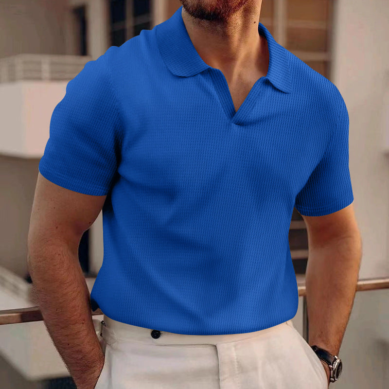 Mann in blauem Polohemd und weißer Hose auf Balkon, modernes Stadtgebäude im Hintergrund. Elegante Herrenmode, Sommeroutfit, urbane Kulisse.