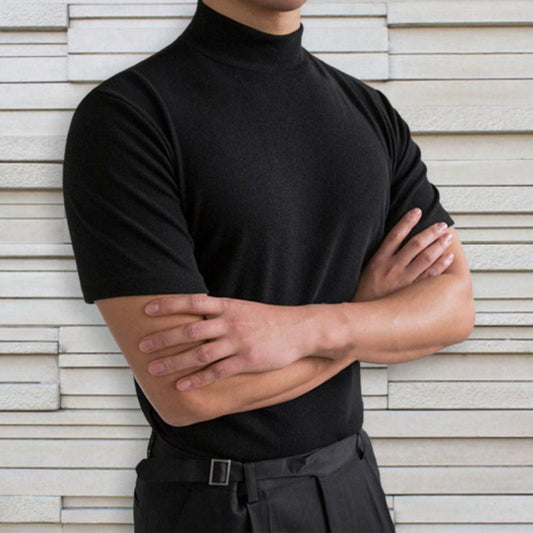 Mann in schwarzem Rollkragenpullover vor weißer Ziegelwand, Arme verschränkt. Elegante Herrenmode, minimalistischer Stil, zeitloses Design.