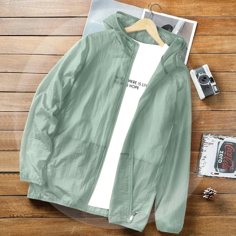 Grüne Windjacke mit Kapuze auf Holztisch, daneben Kamera und Buch. Leichte Herrenjacke, ideal für Outdoor-Aktivitäten und Freizeitmode.