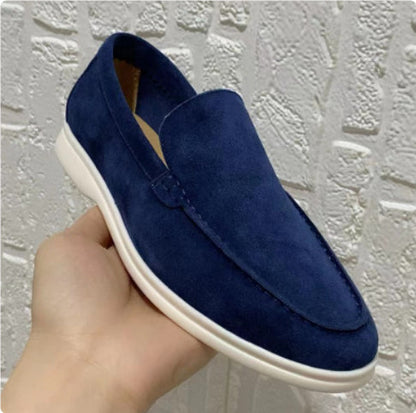 Blaue Wildleder-Slipper für Herren, handgehalten vor weißer Ziegelwand. Stilvolle, bequeme Freizeitschuhe mit weißer Sohle.