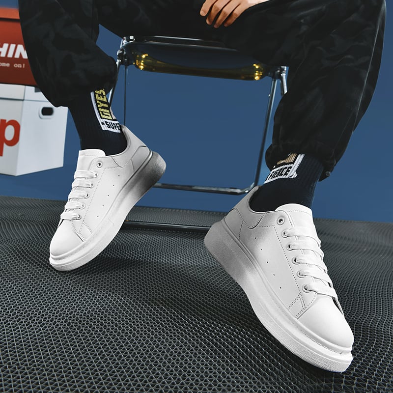 Weiße Sneaker auf schwarzem Metallgitterboden, getragen von Person in schwarzer Hose. Moderne Sportschuhe, urbaner Stil, trendige Modeaccessoires.