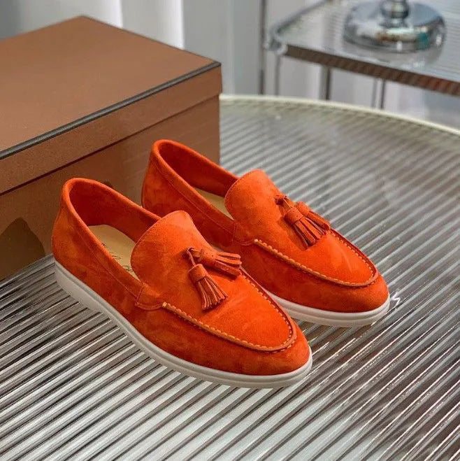 Orange Wildleder-Loafer mit Quasten auf einem gestreiften Tisch, neben einer braunen Box. Modische Herrenschuhe, elegant und modern.