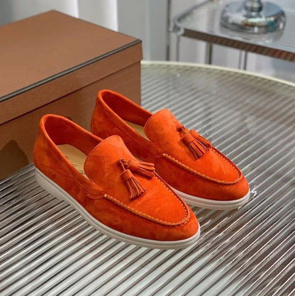 Orange Wildleder-Loafer mit Quasten auf einem gestreiften Tisch, neben einer braunen Box. Modische Herrenschuhe, elegant und modern.