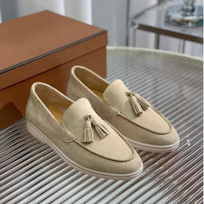 Beige Wildleder-Loafer mit Quasten auf einem silbernen Tisch, neben einer braunen Schuhschachtel. Elegante Herrenschuhe, modisch und stilvoll.