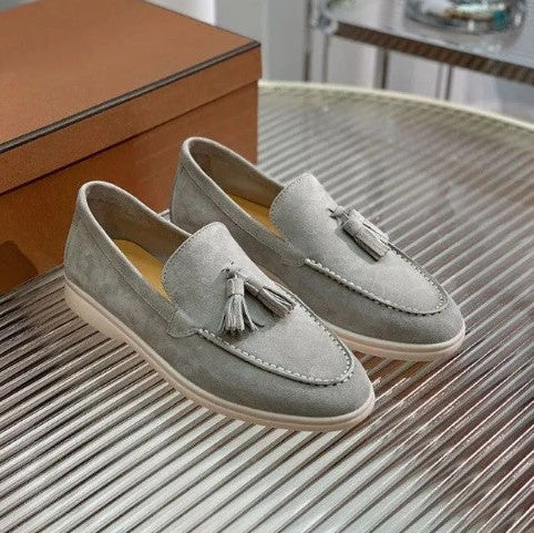 Graue Wildleder-Slipper mit Quasten auf einem geriffelten Tisch, neben einer braunen Schuhschachtel. Elegante Herrenschuhe, modisch und stilvoll.