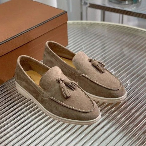 Beige Wildleder-Slipper mit Quasten auf einem Glastisch, elegantes Design, Herrenschuhe, stilvolle Freizeitmode, hochwertige Verarbeitung.