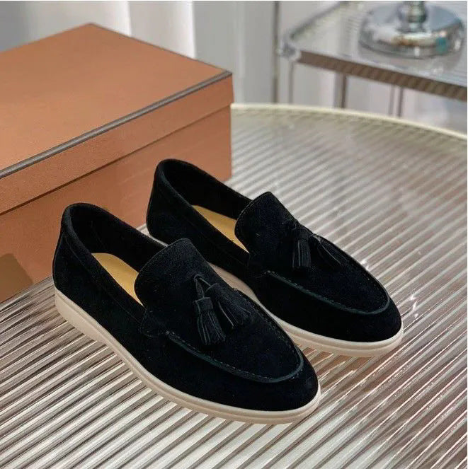 Schwarze Wildleder-Slipper mit Quasten auf einem geriffelten Tisch, daneben eine braune Schuhschachtel. Elegante Herrenschuhe, stilvoll und modern.