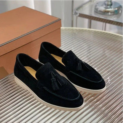 Schwarze Wildleder-Slipper mit Quasten auf einem geriffelten Tisch, daneben eine braune Schuhschachtel. Elegante Herrenschuhe, stilvoll und modern.