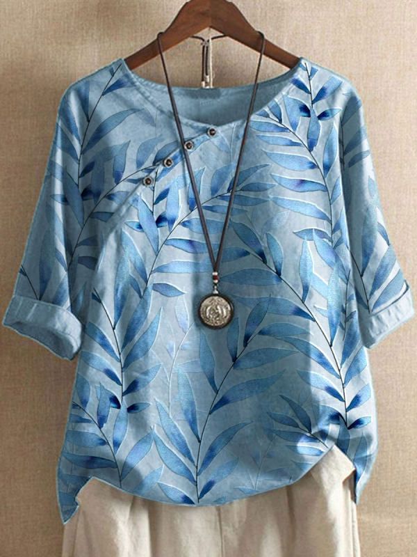 Blaues Damenoberteil mit Blattmuster, asymmetrischem Knopfdesign und Rundhalsausschnitt, kombiniert mit einer langen Halskette und beiger Hose.