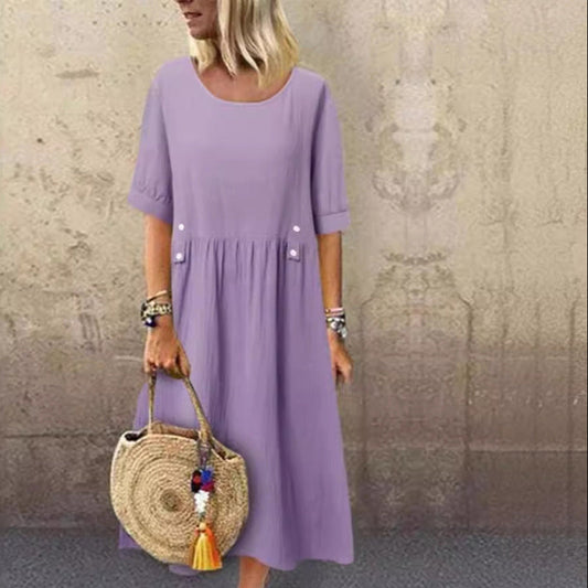 Frau in lila Kleid mit kurzen Ärmeln, hält runde Strohtasche mit bunten Quasten, steht vor beigem Hintergrund. Sommermode, lässiger Stil.