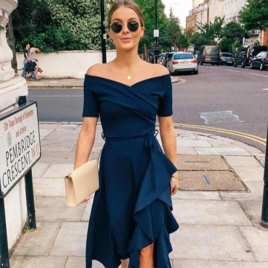 Eine Frau in einem eleganten, dunkelblauen Kleid mit Sonnenbrille und beiger Clutch geht auf einer städtischen Straße. Mode, Sommer, Stadtleben.