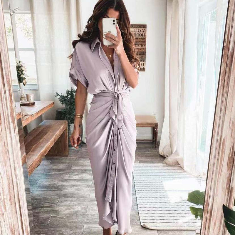 Frau in lila Hemdkleid mit Knoten, steht in modernem Wohnzimmer mit Holztisch und Pflanzen, Spiegel-Selfie. Elegantes, sommerliches Outfit.