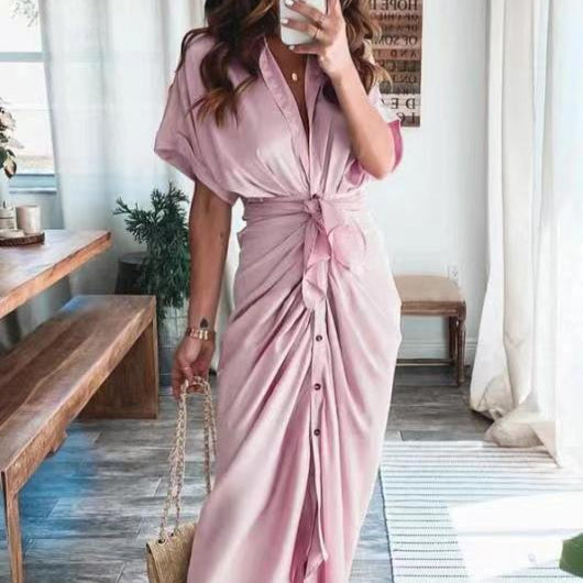 Frau in rosa Maxikleid mit Knopfleiste und Knoten, hält eine beige Handtasche. Steht in modernem, lichtdurchflutetem Raum mit Holzmöbeln.