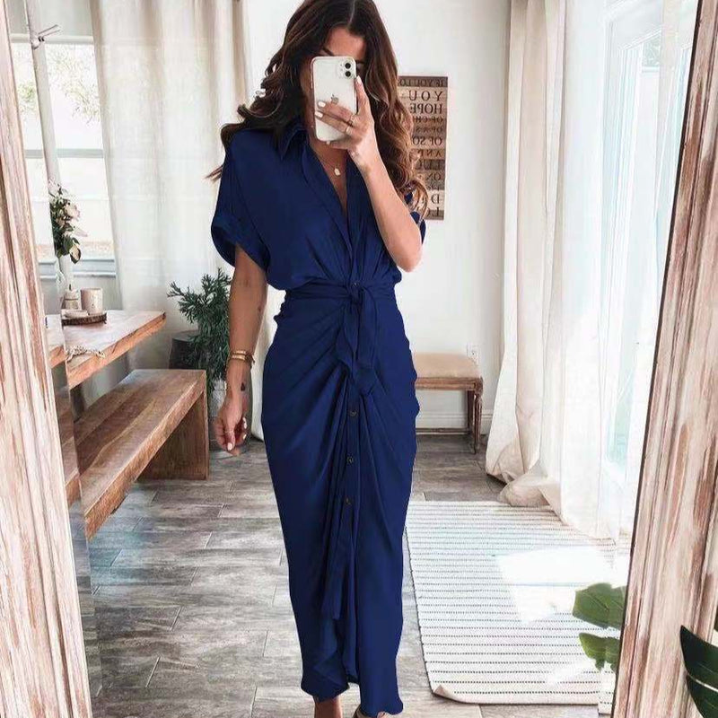 Frau in elegantem, blauem Maxikleid mit Knopfleiste und Taillengürtel, steht in modernem, lichtdurchflutetem Raum mit Holzmöbeln und großen Fenstern.