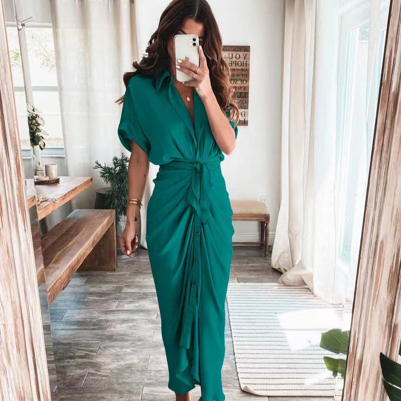 Frau in grünem Kleid mit Knopfleiste und Taillenknoten, steht in modernem Wohnzimmer mit Holztisch und großen Fenstern. Elegante Mode, Innenraum.