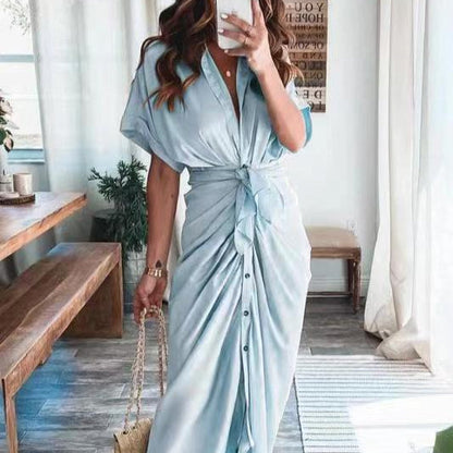 Frau in hellblauem, geknotetem Maxikleid posiert in modernem Wohnzimmer, hält Smartphone und goldene Handtasche. Elegante Mode, Sommerkleid, Innenraum.
