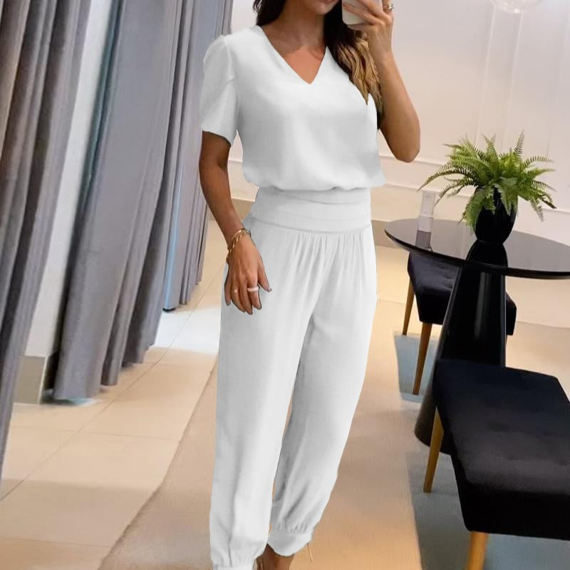 Frau in weißem, lässigem Jumpsuit posiert in modernem Wohnzimmer mit schwarzem Tisch und Pflanze. Elegante, minimalistische Mode, Innenarchitektur.