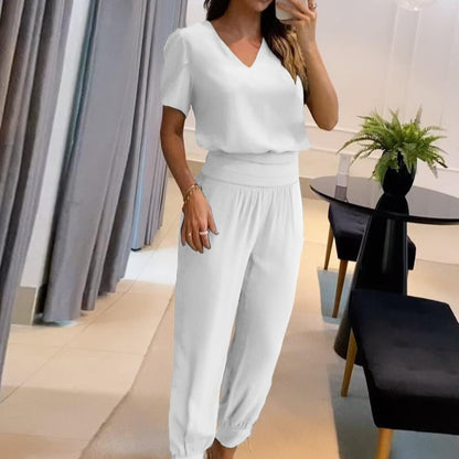 Frau in weißem, lässigem Jumpsuit posiert in modernem Wohnzimmer mit schwarzem Tisch und Pflanze. Elegante, minimalistische Mode, Innenarchitektur.