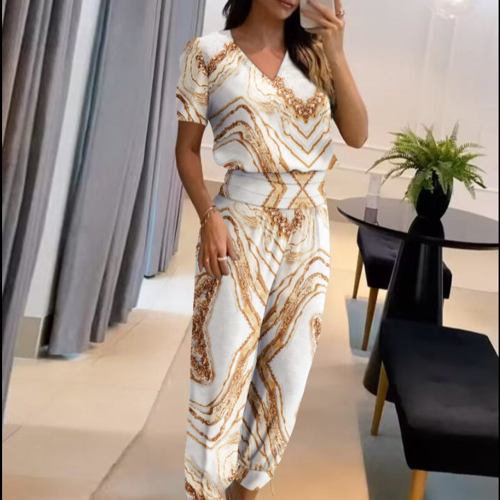 Frau in modernem, weiß-goldenem Jumpsuit mit abstraktem Muster, steht in stilvollem Wohnzimmer mit Pflanzen und Spiegel, modische Damenbekleidung.