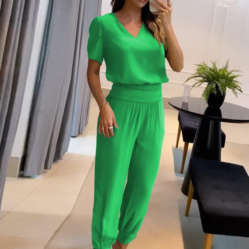 Frau in grünem Jumpsuit steht in modernem Wohnzimmer, elegante Einrichtung, stilvolle Mode, Innenarchitektur, Pflanzen, Vorhänge, Tisch.