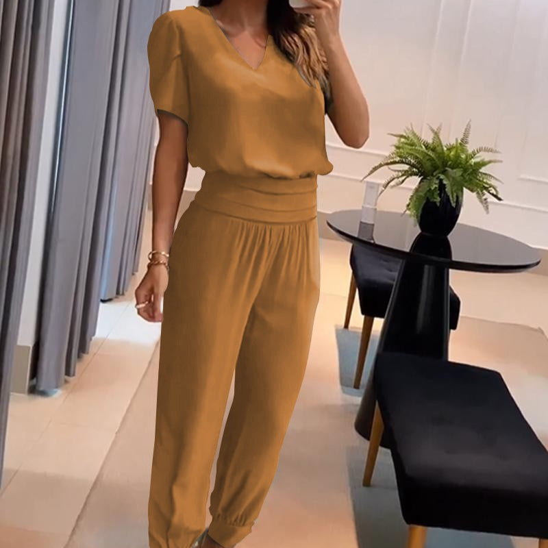 Frau in eleganter, senffarbener Jumpsuit-Mode posiert in modernem Wohnzimmer mit schwarzem Tisch und grüner Pflanze. Trendige Damenbekleidung.
