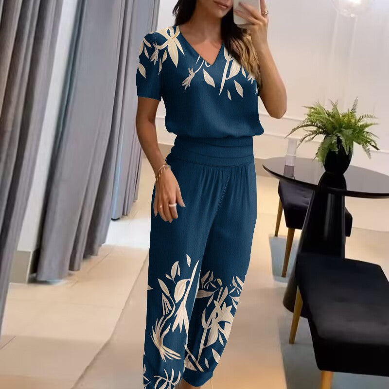 Frau in blauem, geblümtem Jumpsuit macht Selfie; modernes Wohnzimmer, elegante Einrichtung, Pflanzen im Hintergrund. Fashion, Damenmode, Freizeitkleidung.