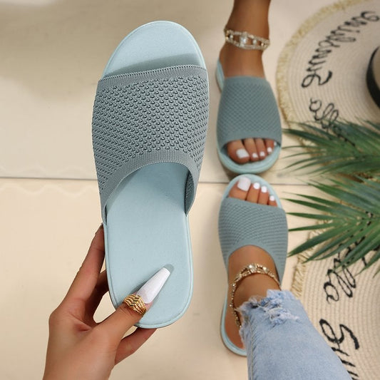 Damenhand hält blaue Strandsandale mit Strickmuster, getragen von Person mit Fußkettchen. Sommermode, bequeme Schuhe, stilvolle Sandalen.