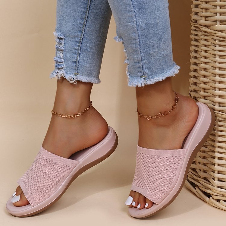 Damenfüße in rosa Strandsandalen mit geflochtenem Design, kombiniert mit zerrissenen Jeans und goldenen Fußkettchen, neben einem Korb.