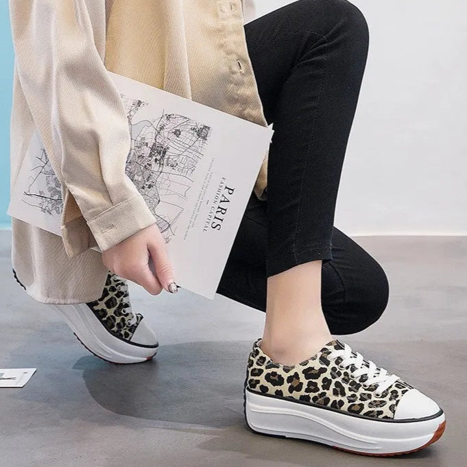 Frau in schwarzer Hose und beigen Hemd hält ein Paris-Poster, trägt Leopardenmuster-Sneaker mit Plateau, sitzt auf grauem Boden.