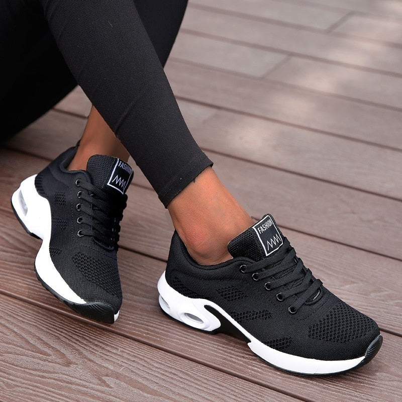 Schwarze Damen-Sneaker mit weißer Sohle, atmungsaktives Mesh-Design, ideal für Sport und Freizeit, auf Holzdeck getragen.
