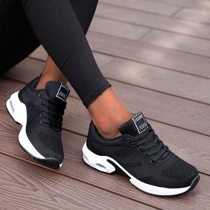 Schwarze Damen-Sneaker mit weißer Sohle, atmungsaktives Mesh-Design, ideal für Sport und Freizeit, auf Holzdeck getragen.