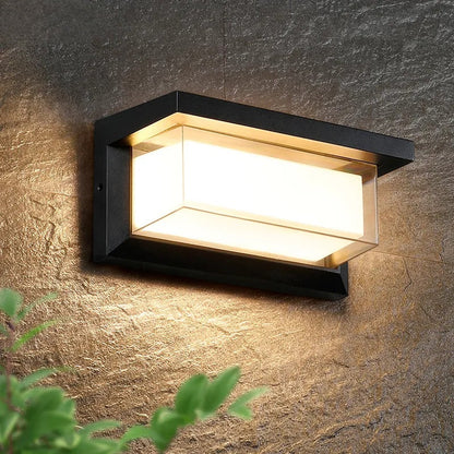 Moderne Außenwandleuchte mit rechteckigem, schwarzem Rahmen und warmweißem Licht, montiert an einer strukturierten Steinwand, umgeben von grünen Blättern.