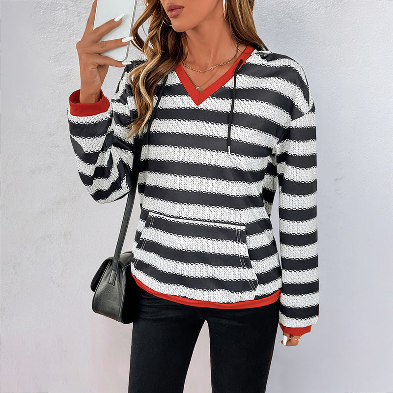 Frau in gestreiftem Pullover mit Kapuze, schwarz-weiß, roter Saum, hält Smartphone, schwarze Tasche, modisches Outfit, lässiger Stil.