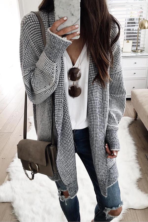 Frau in grauem Strickcardigan, weißem T-Shirt und zerrissenen Jeans, hält Smartphone mit Marmorhülle, braune Umhängetasche, Sonnenbrille.