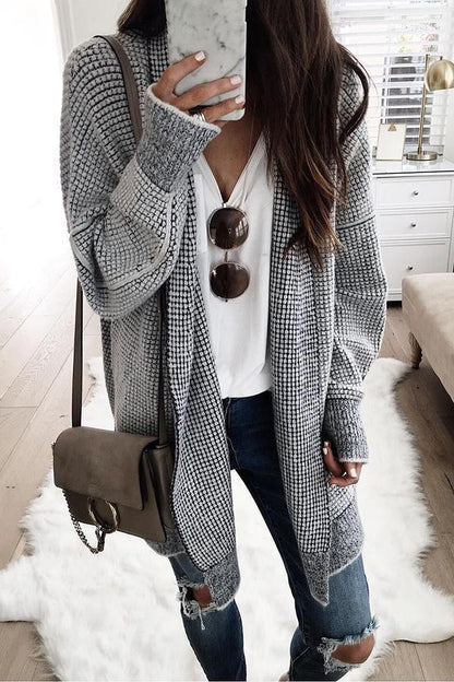 Frau in grauem Strickcardigan, weißem T-Shirt und zerrissenen Jeans, hält Smartphone mit Marmorhülle, braune Umhängetasche, Sonnenbrille.