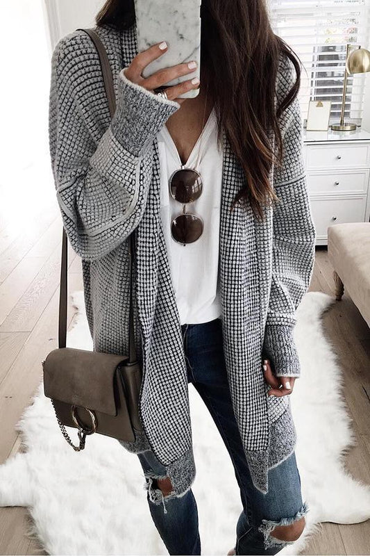 Frau in grauem Strickcardigan, weißem T-Shirt und zerrissenen Jeans, hält Smartphone mit Marmorhülle, braune Umhängetasche, Sonnenbrille.