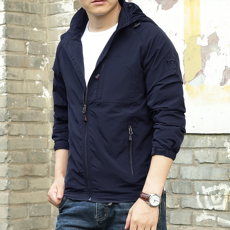 Mann in dunkelblauer Windjacke mit Kapuze, Reißverschlüssen und Armbanduhr, vor einer Backsteinwand. Freizeitmode, Herrenjacke, Outdoor-Bekleidung.