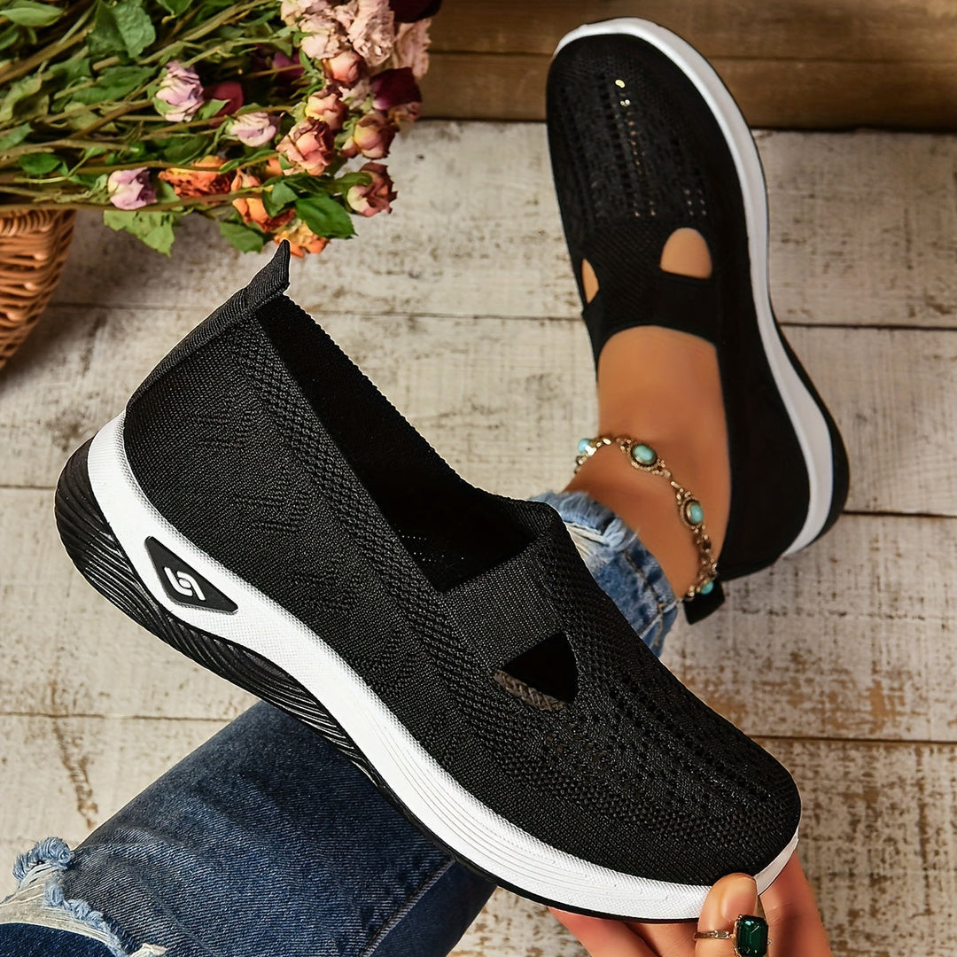 Schwarze Damen-Sneaker mit weißer Sohle, atmungsaktivem Mesh-Obermaterial, getragen von Person mit Jeans und Armband, auf Holzboden.
