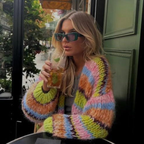 Eine Frau mit blonden Haaren und Sonnenbrille trinkt einen Cocktail in einem Café. Sie trägt eine bunte Strickjacke.