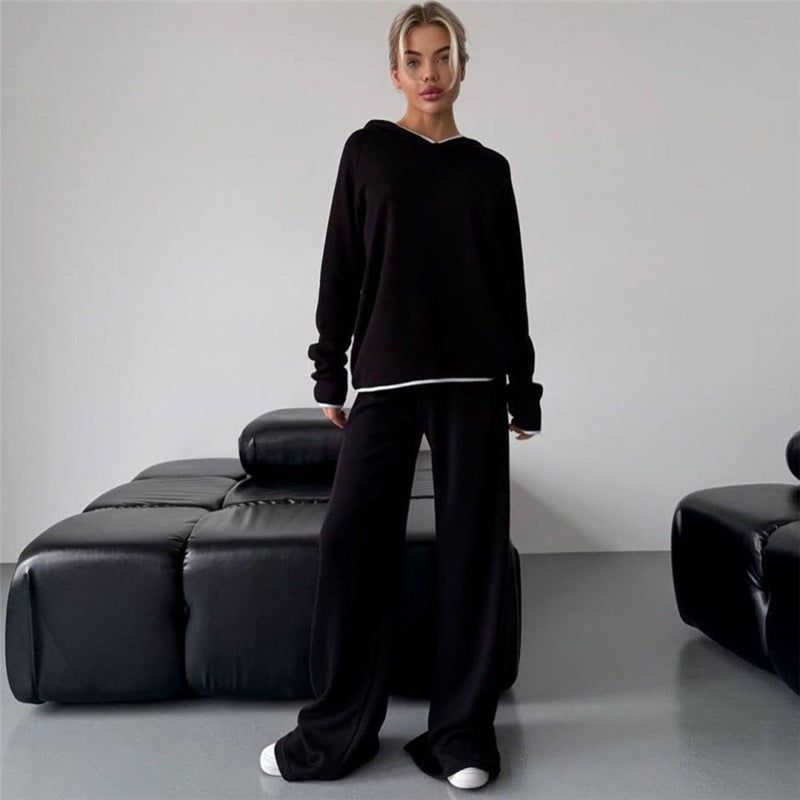 Frau in schwarzem Hoodie und Jogginghose steht vor modernem, schwarzem Ledersofa. Lässige Mode, minimalistische Einrichtung, gemütlicher Look.