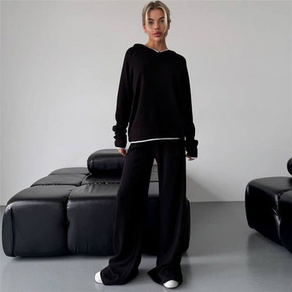 Frau in schwarzem Hoodie und Jogginghose steht vor modernem, schwarzem Ledersofa. Lässige Mode, minimalistische Einrichtung, gemütlicher Look.