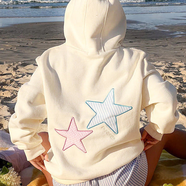 Person in weißem Hoodie mit rosa und blauem Stern-Design sitzt am Strand. Sommermode, Strandbekleidung, Freizeitkleidung, Hoodie mit Sternen.
