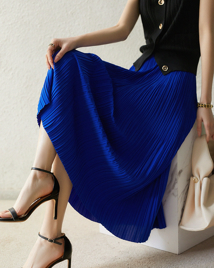 Frau in blauem Plisseerock und schwarzem Oberteil, sitzt auf einem Stuhl. Elegante schwarze Sandalen, modisches Outfit, stilvolle Damenmode.